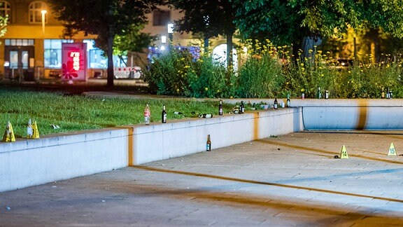 Auf einer Betonmauer stehen Flaschen und Polizeischilder zur Markierung von Spuren.