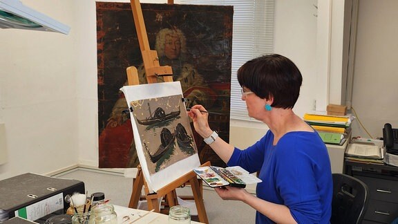 Eine Frau mit kurzen, dunklen Haaren, Brille und dunkelblauer Bluse sitzt mit Pinsel und Farbpalette vor einem historischen Gemälde auf einer Staffelei in einem Atelier.