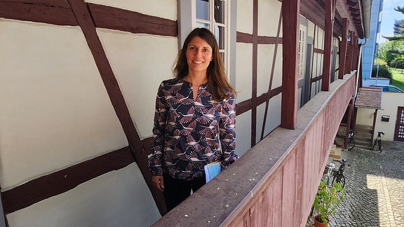 Eine Frau mit langen, offenen, braunen Haaren und bunter Bluse steht im Laubengang eines alten Fachwerkhauses und lächelt in die Kamera.