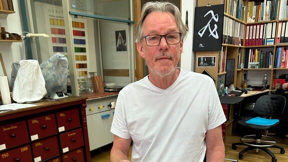 Ein Mann mit mittellangen, grauen Haaren, Brille, weißem T-Shirt und Jeans sitzt in einem Atelier hinter einer aufrecht gestellten, historischen Holztruhe und hält einen Pinsel in der Hand.
