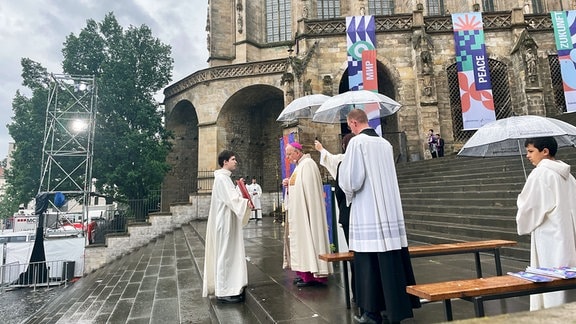 Der Erfurter Bischof Ulrich Neymeyr steht auf den Domstufen und ließt etwas vor. Neben ihm Stehen Messdienser die Regenschirme halten. Einer hält eine Bibel.