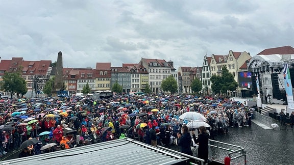 Viele Menschen stehen auf dem Domplatz. Weil es Regenet, tragen sie Regenjacken oder halten Schirme in der Hand.
