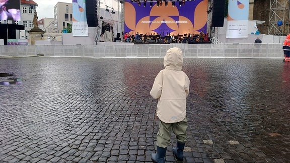 Ein Kind steht in Regenkleidung auf dem nahezu leeren Domplatz in Erfurt vor der Bühne für den Katholikentag.