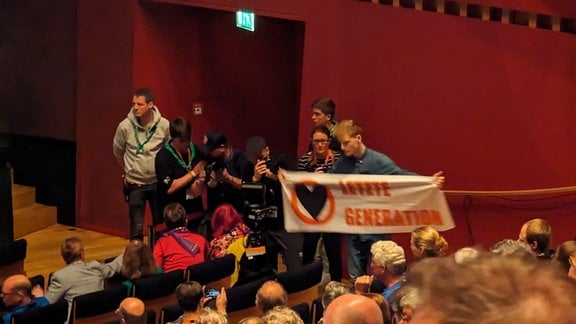 Eine Gruppe Klimaaktivisten steht am Rand des Theatersaals in Erfurt und halten ein Banner mit der Aufschrift "Letzte Generation" vor sich.