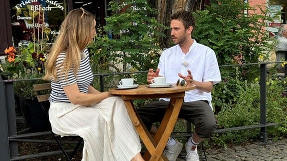 Sänger Clueso sitzt mit einer Frau an einem Tisch im Außenbereich eines Kaffees