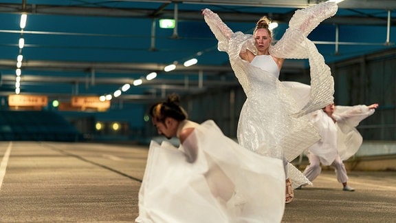 Drei Personen tanzen im Rahmen einer Perfomance in weißer Plastikfolie gehüllt in einer Halle