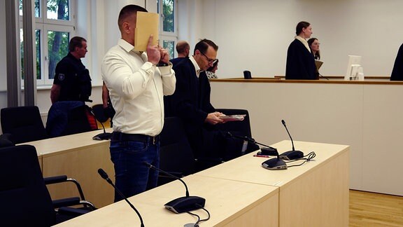 Ein Mann hält sich in einem Gerichtssaal einen Ordner vor sein Gesicht.