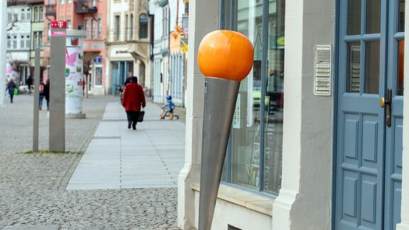 Ein schmaler auf dem Kopf stehender Metallkegel. Darauf eine große, orange Kugel. Die Nadel steht vor einem Geschäft.
