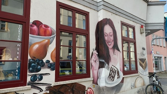 Eine kunstvoll bemalte Häuserfassade zeigt eine lächelnde Frau, Kaffe und Obst. 