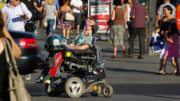 Ein junger Mann im Rollstuhl mit Beatmungsmaske im Stadtverkehr. Im Vordergrund ein Auto, ringsum Passanten. 