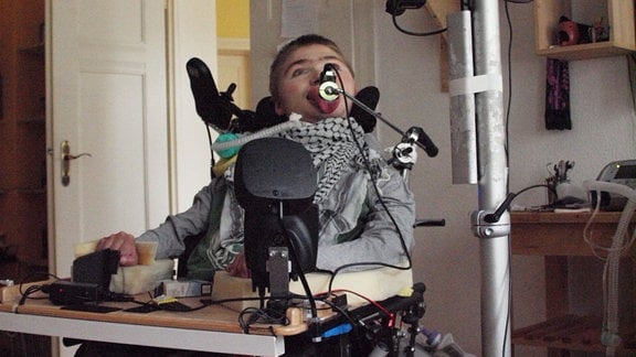 Ein junger Mann im Rollstuhl, davor ein Computer. Ein Schlauch führt vom Beatmungsgerät zu seinem Hals (Trachealkanüle). 