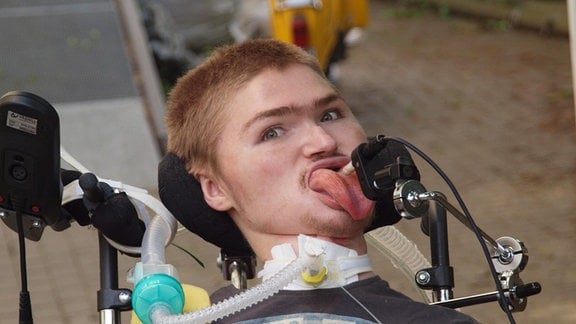 Ein junger Mann im Rollstuhl. Vor seinem Gesicht ein Joystick, den er mit seiner Zunge steuert. Ein Schlauch führt vom Beatmungsgerät zu seinem Hals. 