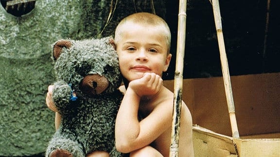 Ein Junge oberkörperfrei mit Sommershorts und Sandalen. Im Arm hält er einen graubraunen Teddybär. 