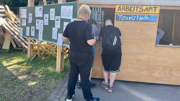 Zwei Jungs stehen an einer Holzhütte. Daran hängen die Schilder "Arbeitsamt" und "Touristinfo".
