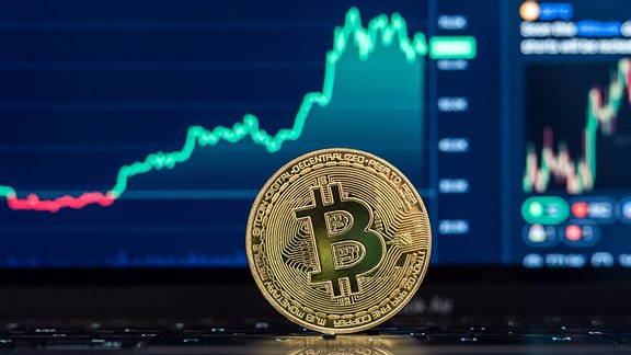 Das Logo der Kryptowährung Bitcoin BTC ist auf einer Münze zu sehen, die vor einem Bitcoinchart der Handelsplattform.