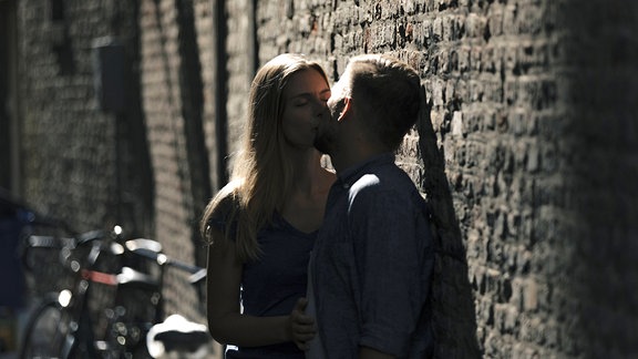 Junger Mann und junge Frau küssen sich an einer Mauer.