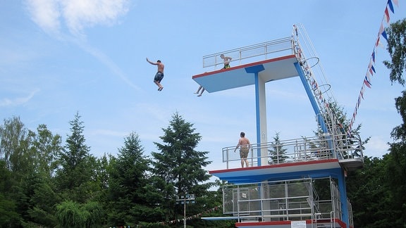 Mann springt von 10m-Turm in Sprunganlage mit drei weiteren Sprungbretthöhen in  einem von Bäumen umstandenen Freibad. Zuschauer am Beckenrand und anderen Brettern.