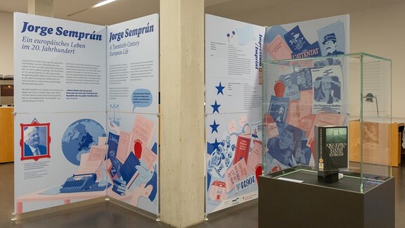 Große Aufsteller mit blauem Text auf weißem Hintergrund und blau-roten Illustrationen stehen in einer Ausstellung.