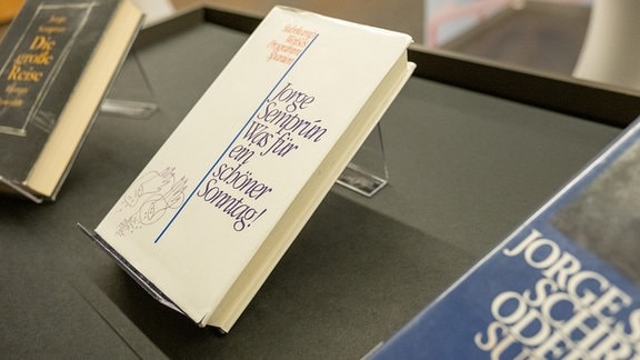 Bücher in einer Ausstellung, in der Mitte ein Buch im weißen Einband mit der Aufschrift "Jorge Semprún: Was für ein schöner Sonntag!"