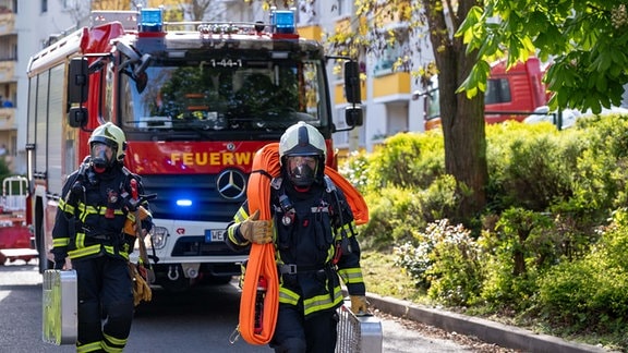 Zwei Feuerwehrmänner tragen Ausrüstung, dahinter ein Feuerwehrfahrzeug.