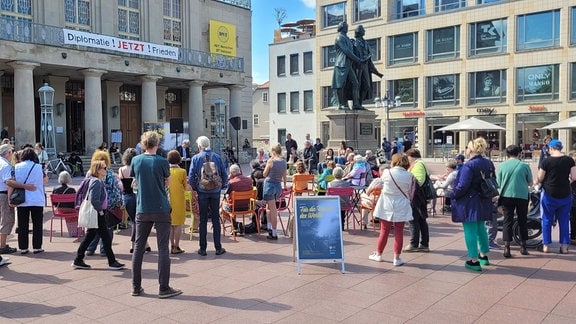 Eine Menschenmenge steht vor dem Deutschen Nationaltheater in Weimar um eine kleine Bühne auf der eine Person etwas vorliest. Auch Tische und Stühle sind aufgestellt.