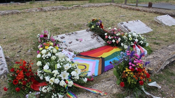 Blumen und eine LGBTQI-Flagge liegen rund um einen Gedenkstein.