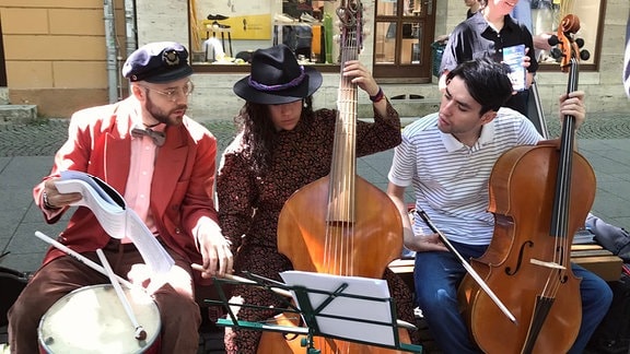 Drei junge Musiker sitzen mit verschiedenen Instrumenten am Straßenrand.