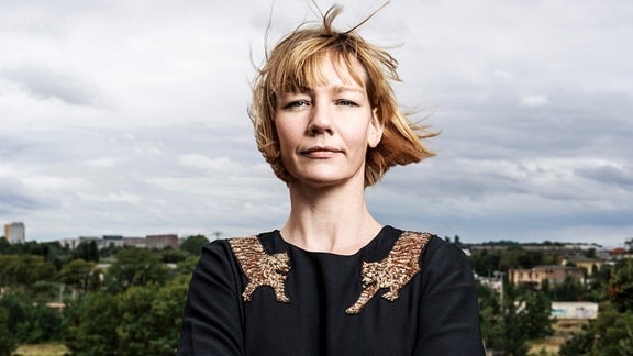 Sandra Hüller, eine Frau mit kinnlangen, vom Wind durchwehten blonden Haaren und schwarzem Oberteil steht vor einer Landschaft und blickt entschlossen in die Kamera.