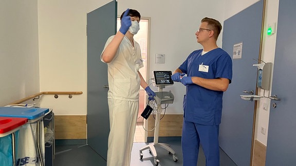 Pfleger und Pflegeschüler stehen mit einem medizinischen Gerät zur Messung von Vitalfunktionen in einem Patientenzimmer.