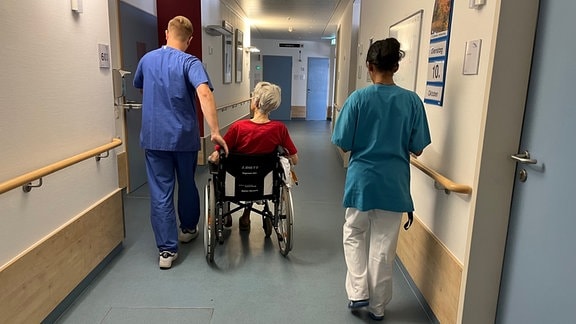 Eine Pflegerin und ein Pfleger schieben eine Patienten in ihrem Rollstuhl durch den Krankenhausgang.