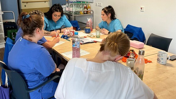 Fünf Pflegerinnen sitzen an einem Tisch und schreiben teilweise etwas auf das bedruckte Blatt, das jeweils vor ihnen liegt.