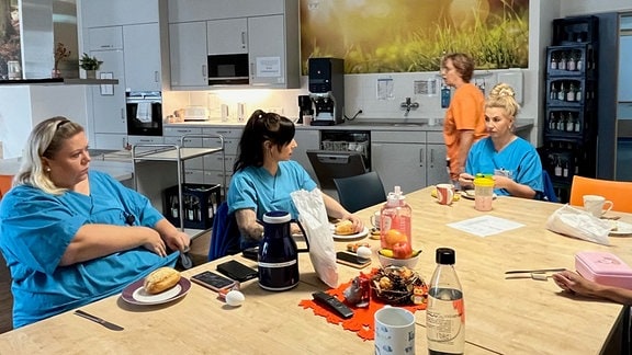 Drei Pflegerinnen sitzen an einem Tisch und haben Teller mit Brötchen vor sich. Auf dem Tisch steht außerdem eine Kaffeekanne, Kaffeetassen, Obst und Wasserflaschen.