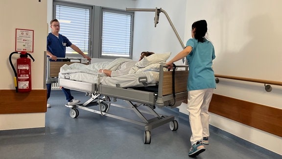 Eine Pflegerin und ein Pfleger schieben eine Patientin in ihrem Bett auf dem Krankenhausgang.
