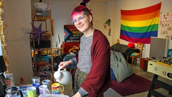 eine Frau mit lila haaren in einem Raum (Beratungsstelle) mit vielen Deko-Elementen in Regenbogenfarben, Flyer und ein Plakat vom Queeren Weihnachtsprogramm, eine Drag-Queen auf einem Stuhl, die einen Monitor anschaut