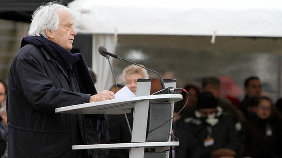 Ein älterer Mann mit weißem Haar und schwarzem Mantel steht an einem Rednerpult.