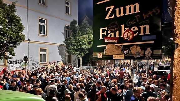 Zahlreiche Menschen stehen auf einer Straße im Dunkeln vor dem Eingang eines Lokals. 