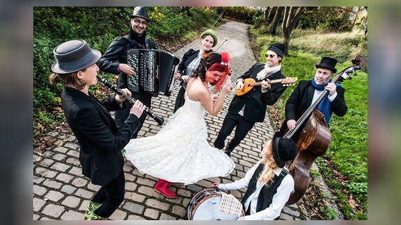 Klezmer-Band "She'Koyokh" aus Großbritannien: Sechs Musiker stehen im Kreis, eine Frau im weißen Kleid tanz in ihrer Mitte