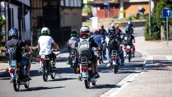 Minschen mit Simson Mopeds fahren durch eine Ortschaft.