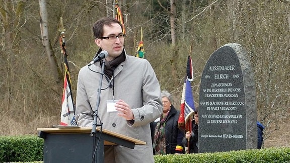 Mann in grauem Mantel an Pult mit Mikrofon. Im Hintergrund ein grauer Obelisk mit weißen Inschriften