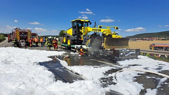  Ein Laster mit einem geladenen Bagger steht auf einer Autobahn. Auf der Fahrbahn ist viel Lösch-Schaum von der Feuerwehr