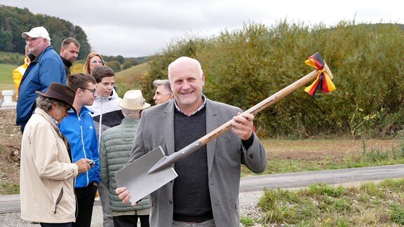 Ein mittelalter Mann - Torsten Städler, der Bürgermeister von Kallmerode - hält einen Spaten mit einer Schleife in den Händen und lacht in die Kamera, daneben stehen einige Personen.