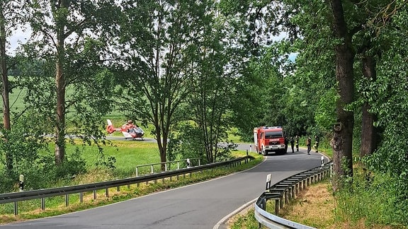 Ein Feuerwehrauto und ein Hubschrauber stehen auf einer bewaldeten Straße.