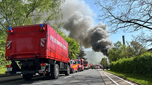 Feuerwehrautos stehen in einer Straße neben einem Fabrikgebäude aus dem eine riesige Rauchwolke steigt. 