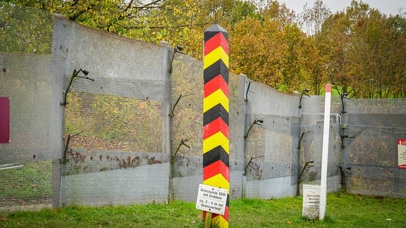 Vor einem mehrere Meter hohen Zaun steht eine schwarz-rot-gelbe Betonsäule mit der Aufschrift "Grenzsäule DDR mit Emblem".