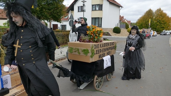 Ein Totengräber zieht einen Pappsarg hinter sich her auf dem steht: "Die Kassen sind leer, ein Pappsarg muss her!"