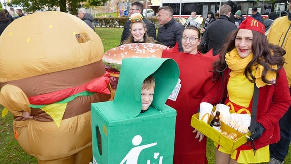 Mehrere Personen bei einem Kirmes-Festumzug, unter anderem verkleidet als Burger und Verkäuferin von McDonalds. Im Vordergrund ist ein Kind als Mülleimer verkleidet.