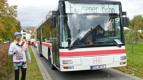 Ein Bus auf einer Straße auf dem "Hausener Bahn" oben angezeigt wird. Daneben läuft ein Mann mit Fischerhut und Getränk in der Hand.