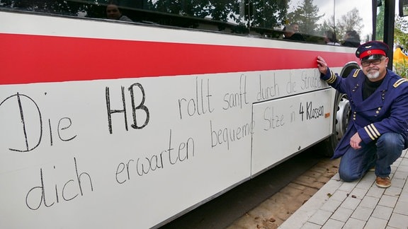 Ein Mann mit Schaffner-Uniform kniet neben einem Bus. Auf dem Bus steht geschrieben: "Der HB rollt sanft durch die Straßen, dich erwarten Sitze in 4 Klassen."