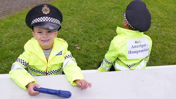 Zwei Kinder in englischen Polizeiuniformen. Sie tragen Hüte. Ein Kind hat ein Schild auf dem Rücken auf dem steht "Mini-Anzeigenhauptmeister"