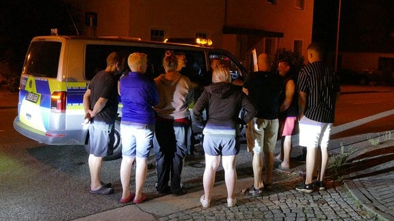 Mehrere Menschen stehen nacht vor einem Polizeiauto.